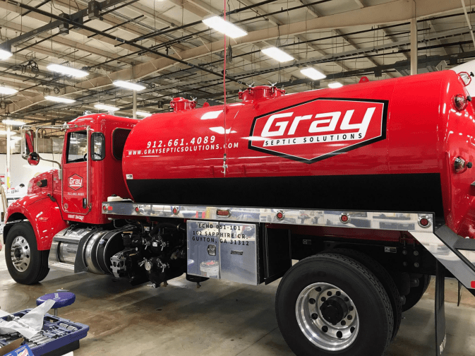 A red Grau Septic Solutions truck is parked inside a warehouse, displaying company branding, contact number, and website on the side of the tank.