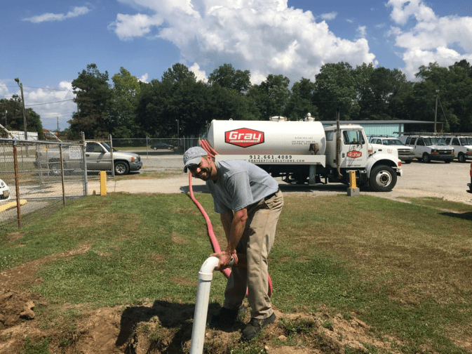 A worker connects a pink hose to a white pipe outdoors, with a Gray company truck and several vehicles in the background.