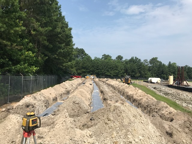 A construction site with long trenches dug in the ground, lined with black material, likely prepared for commercial septic services, surrounded by dirt mounds, trees, and construction equipment under a partly cloudy sky.