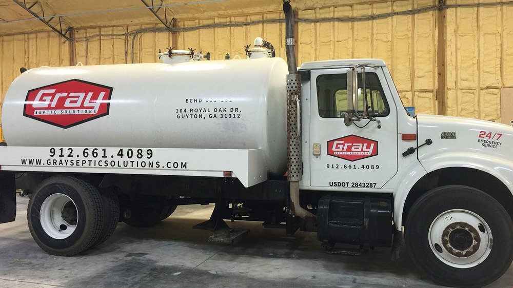 A white Gray Septic Solutions truck is parked indoors, featuring company logo, contact information, and service details on the side of the tank and cab.