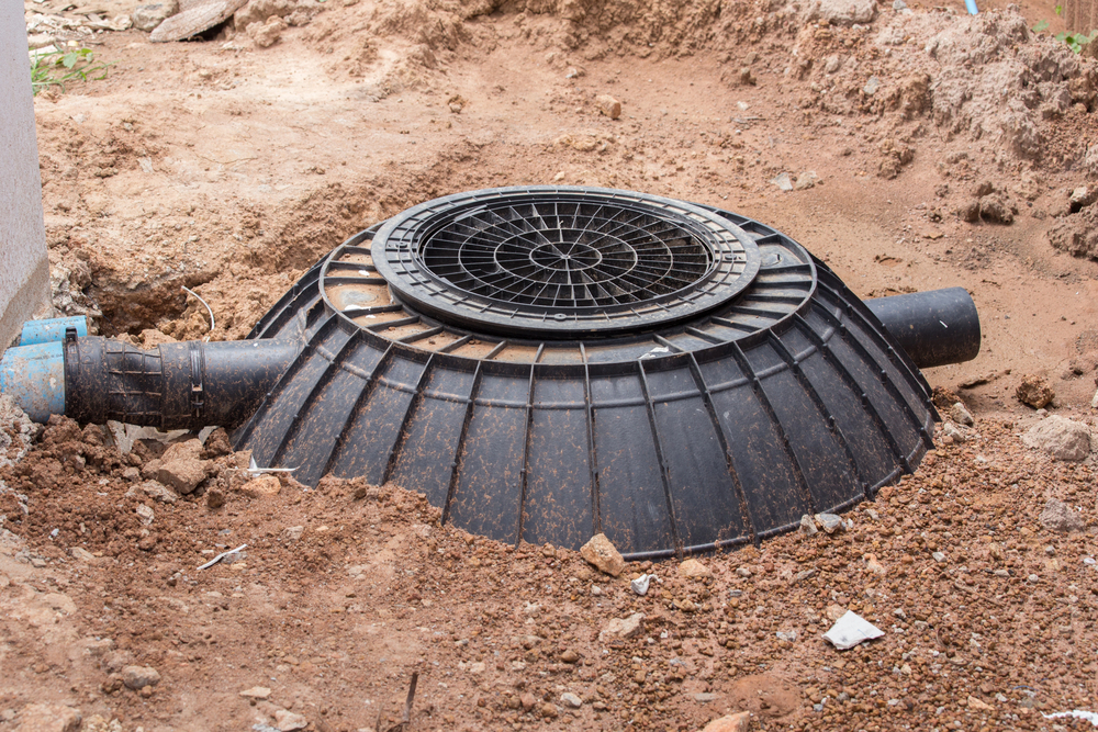 Large black plastic septic tank cover with attached pipes partially buried in brown soil at a construction site.