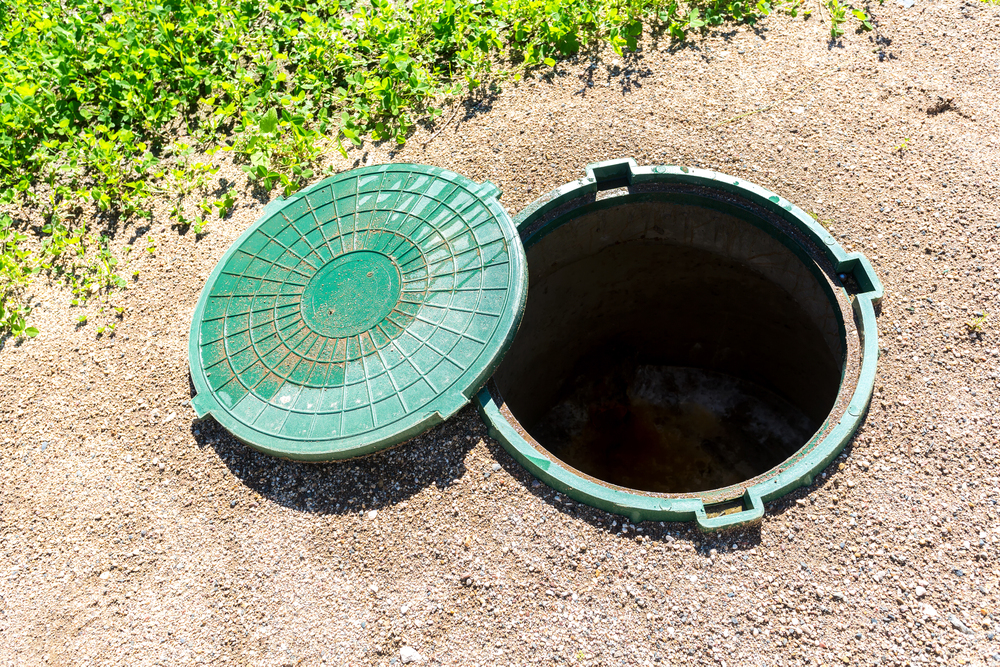 A green manhole cover is open, revealing a circular underground access hole surrounded by dirt and grass.