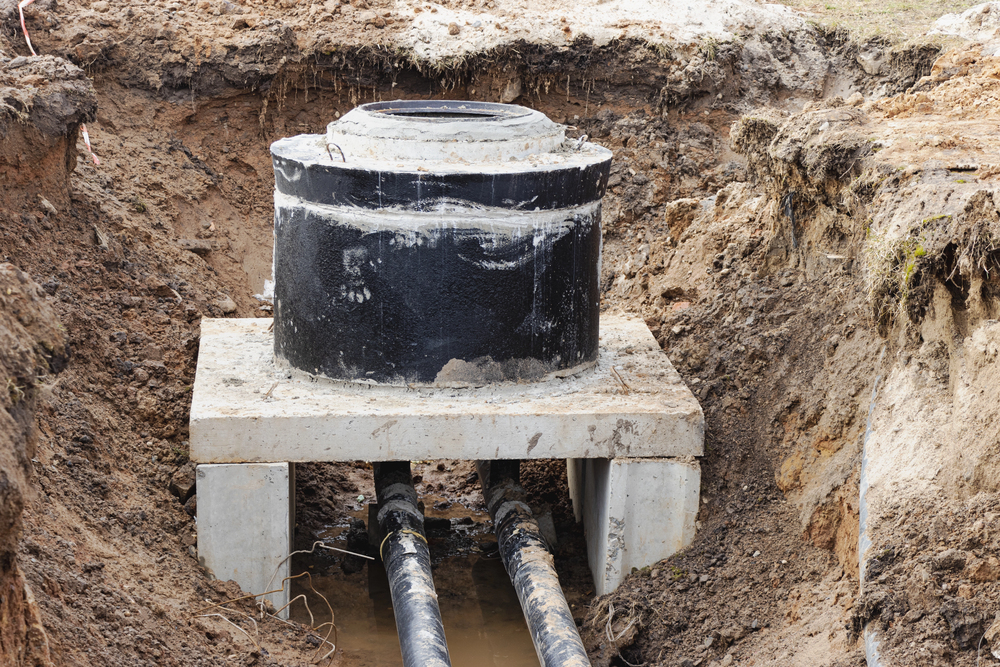 A large black utility vault with two pipes leading into it is installed in an excavated pit surrounded by soil.