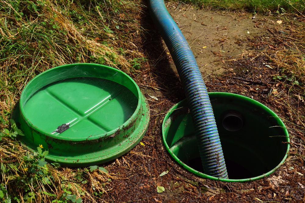 An open green septic tank with its lid off and a large blue hose inserted for pumping or cleaning. The surrounding ground is grassy and uneven.