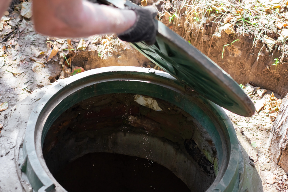 A person in a black glove opens the lid of a large outdoor well or manhole, surrounded by soil and fallen leaves—possibly preparing for a septic tank inspection.