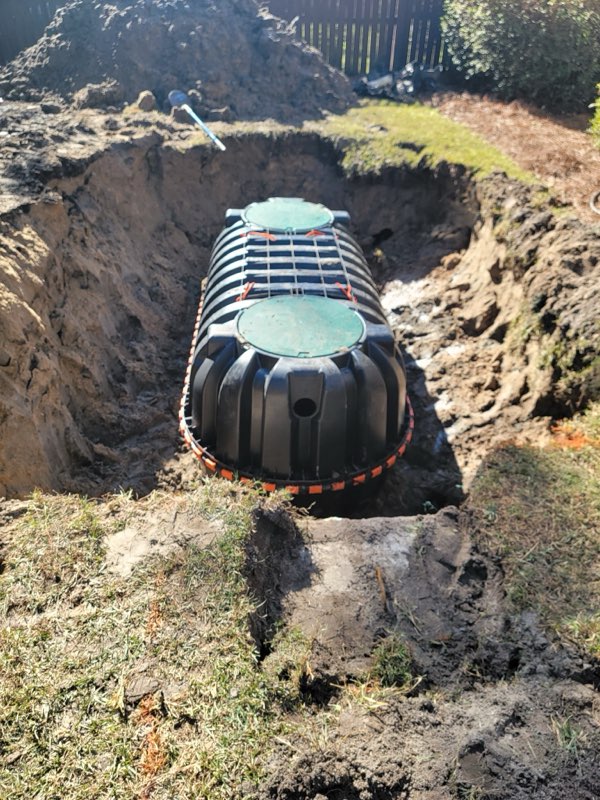 A large black septic tank, partway buried in a dug hole for septic tank installation, sits in a backyard surrounded by dirt and patches of grass.