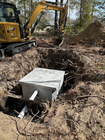 A construction site with a CAT excavator beside an open pit, where a concrete septic tank is being installed underground with visible pipes attached&mdash;part of a comprehensive septic tank repair project.