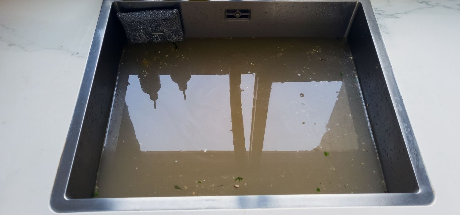 A stainless steel kitchen sink filled with murky, dirty water and food debris&mdash;one of the common septic system problems and warning signs indicating a possible drainage issue.